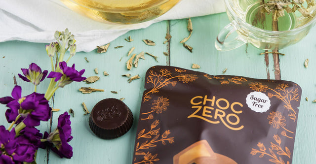 Bag of ChocZero PB Cups on a green wooden table with flowers and loose leaf tea scattered.
